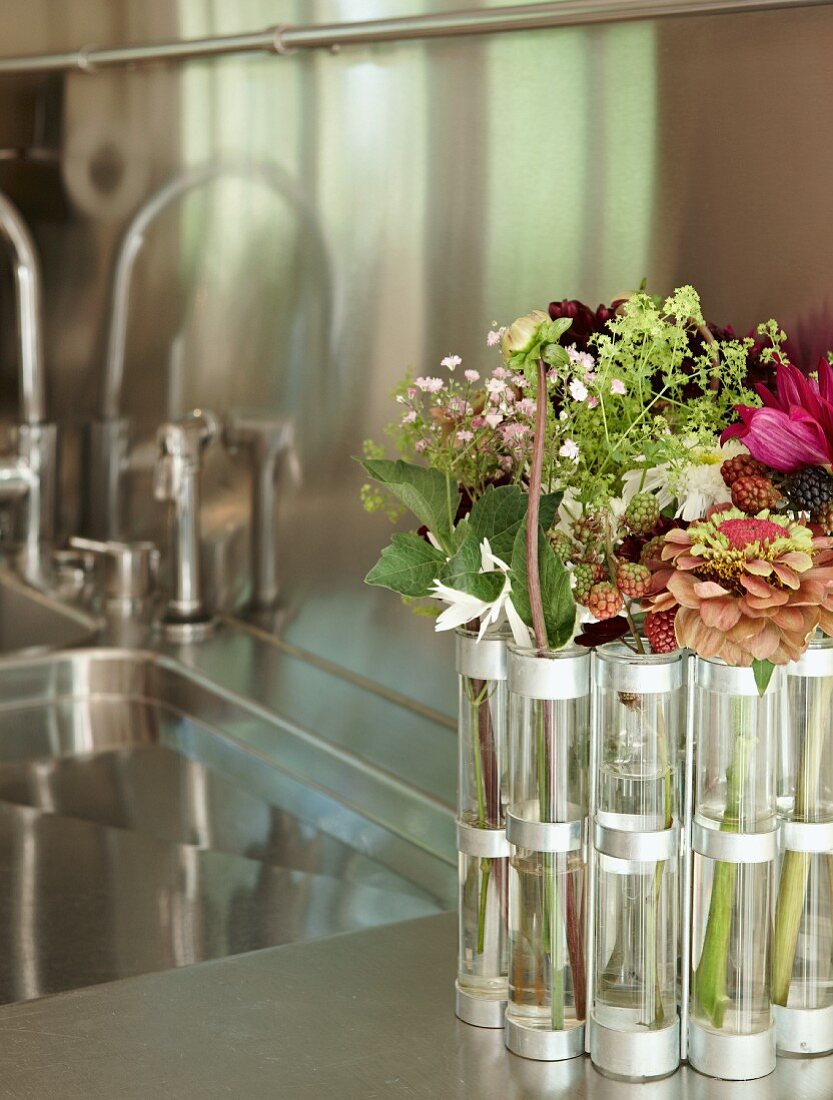 Garden flowers in narrow cylindrical vases next to a kitchen sink in a stainless steel countertop with mirror effect