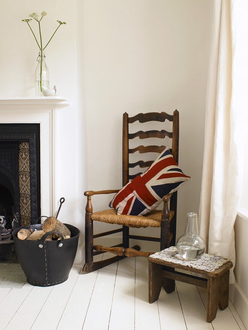 Kissen mit Union Jack Bezug auf altem Schaukelstuhl, Holzschemel und Korb mit Brennholz am Kamin