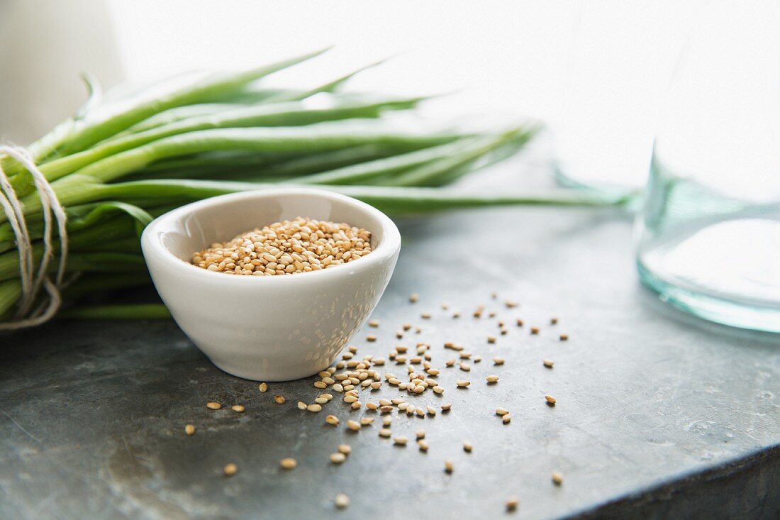 A Bowl of Sesame Seeds with Some Spilled; Green Onions in Background