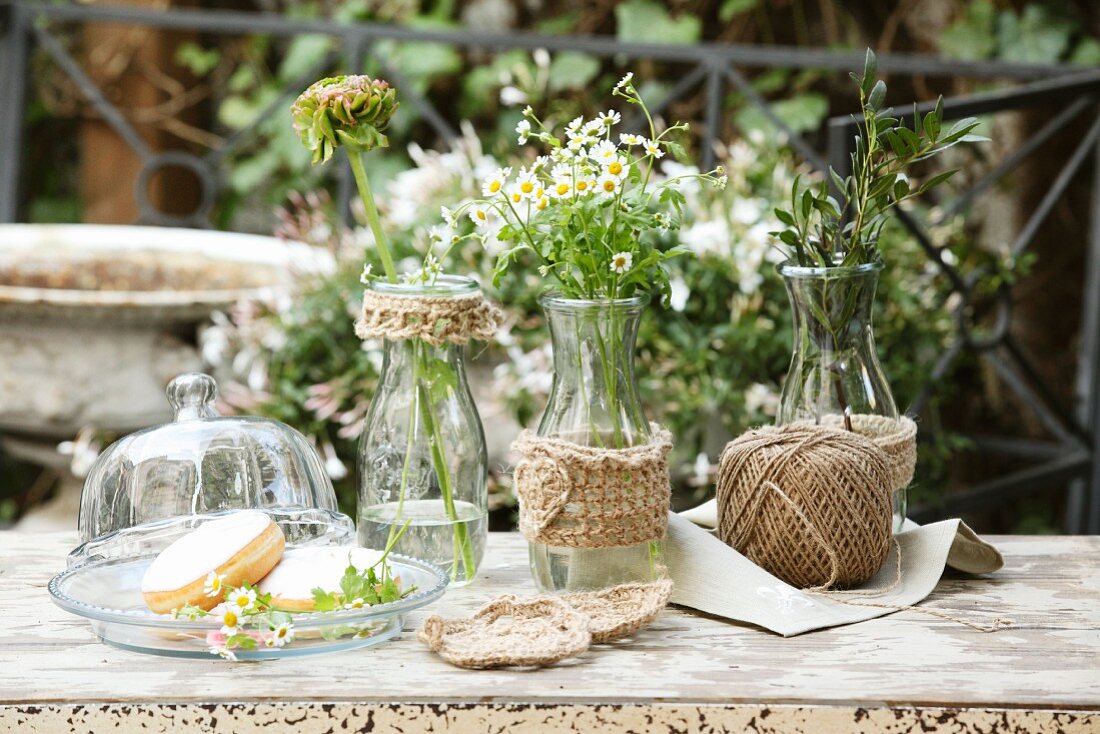 Selbstgestrickte Hüllen für Glasvasen neben Glasglocke auf Gartentisch im Freien