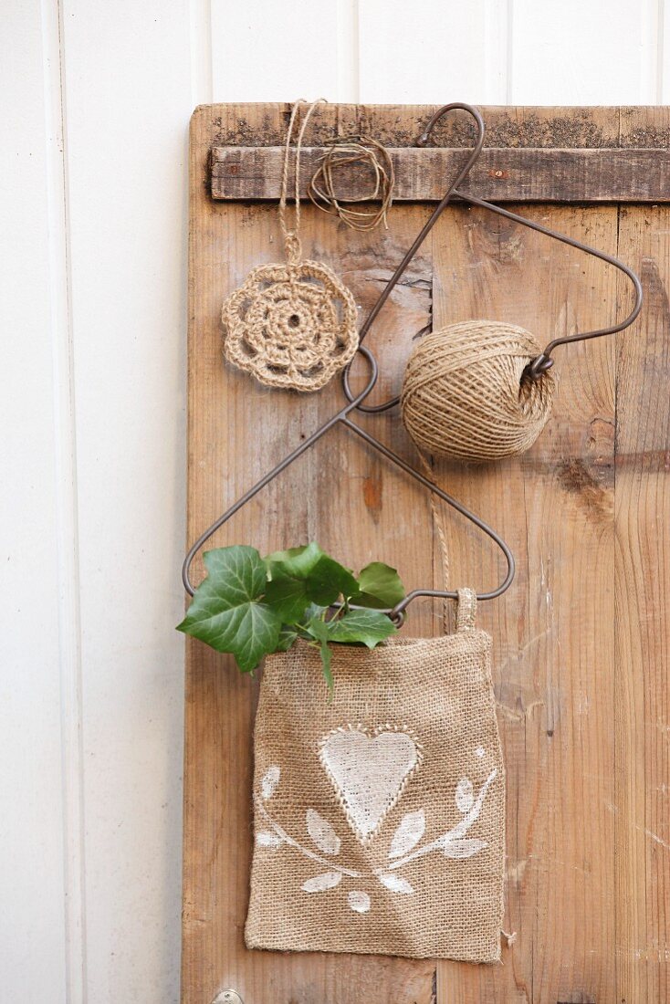 Jute yarn reel and white-painted sacking bag hanging from wire hangers on old wooden panel next to doily crocheted from jute yarn