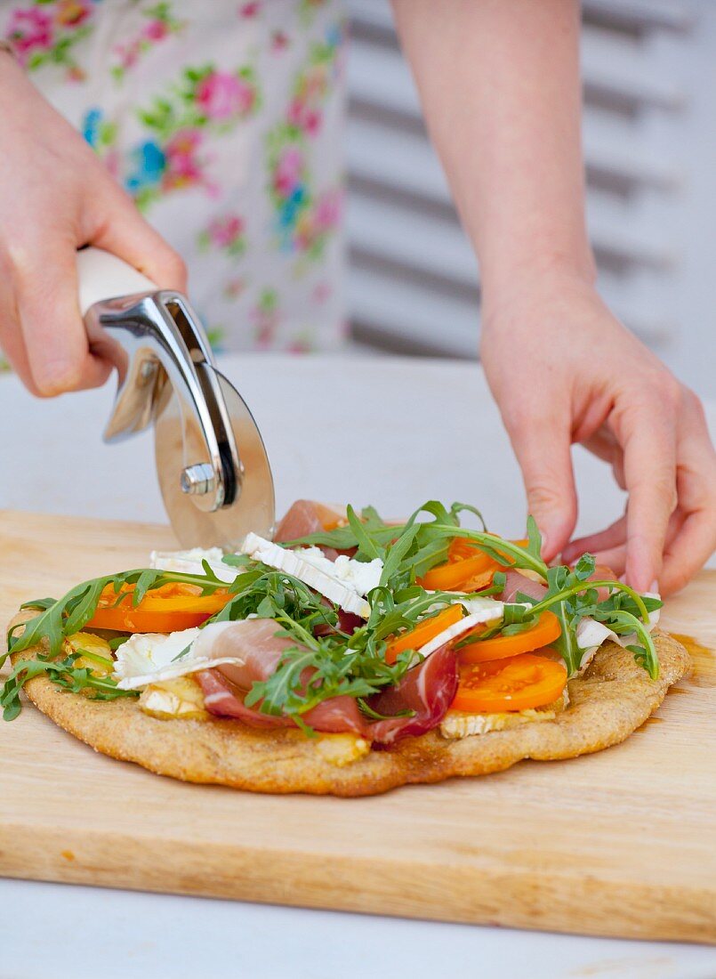 A woman cutting in half a pizza topped with rocket, prosciutto, goat's cheese and tomatoes