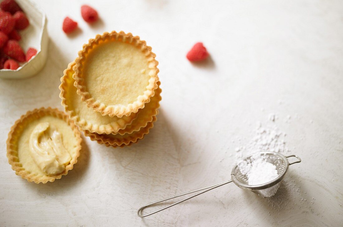 Fertig gebackene Torteletts zum Befüllen, daneben Himbeeren & Puderzucker