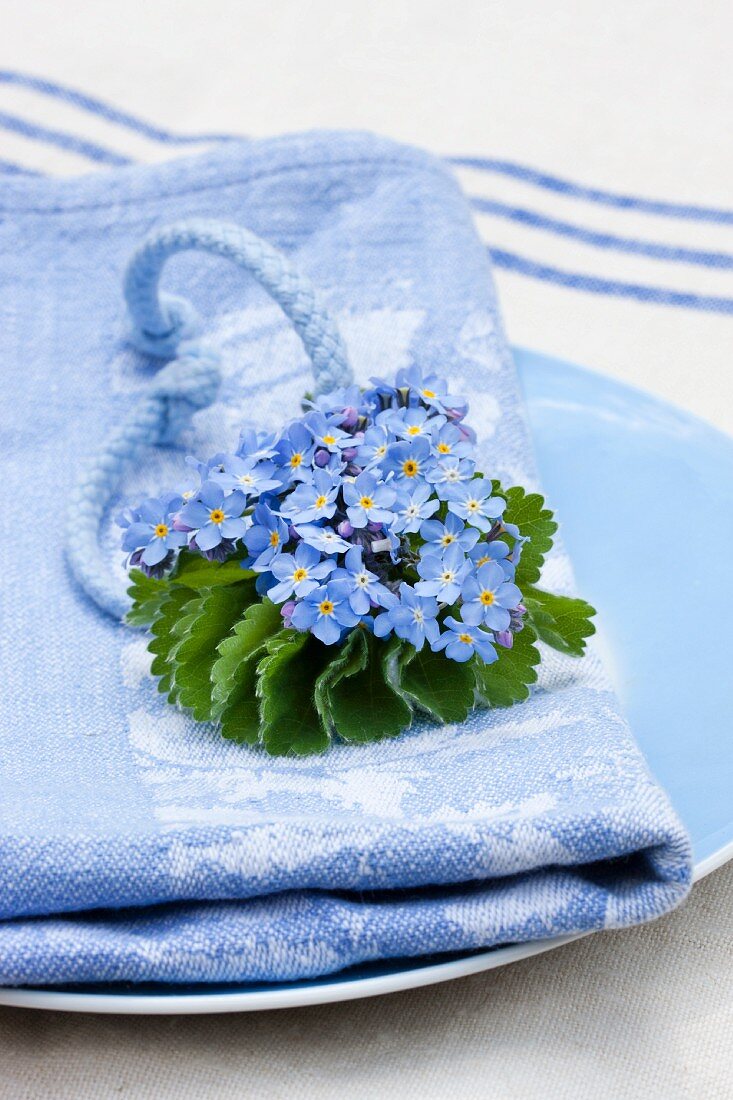 Serviettendeko: Vergissmeinnicht und Frauenmantelblatt mit blauer Kordel auf blauer Serviette, darunter Leinentuch