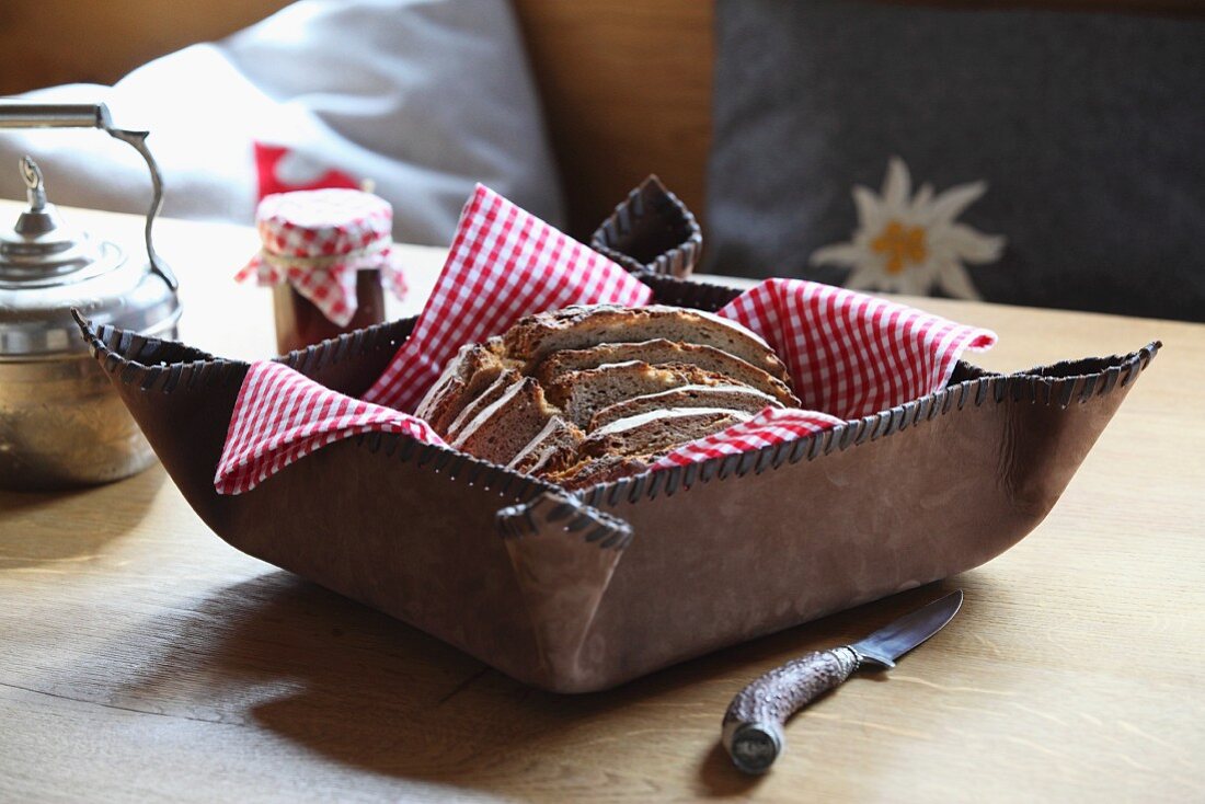 Rustikaler Brotkorb aus umnähtem Leder mit rotweisskarierter Serviette und Brotscheiben