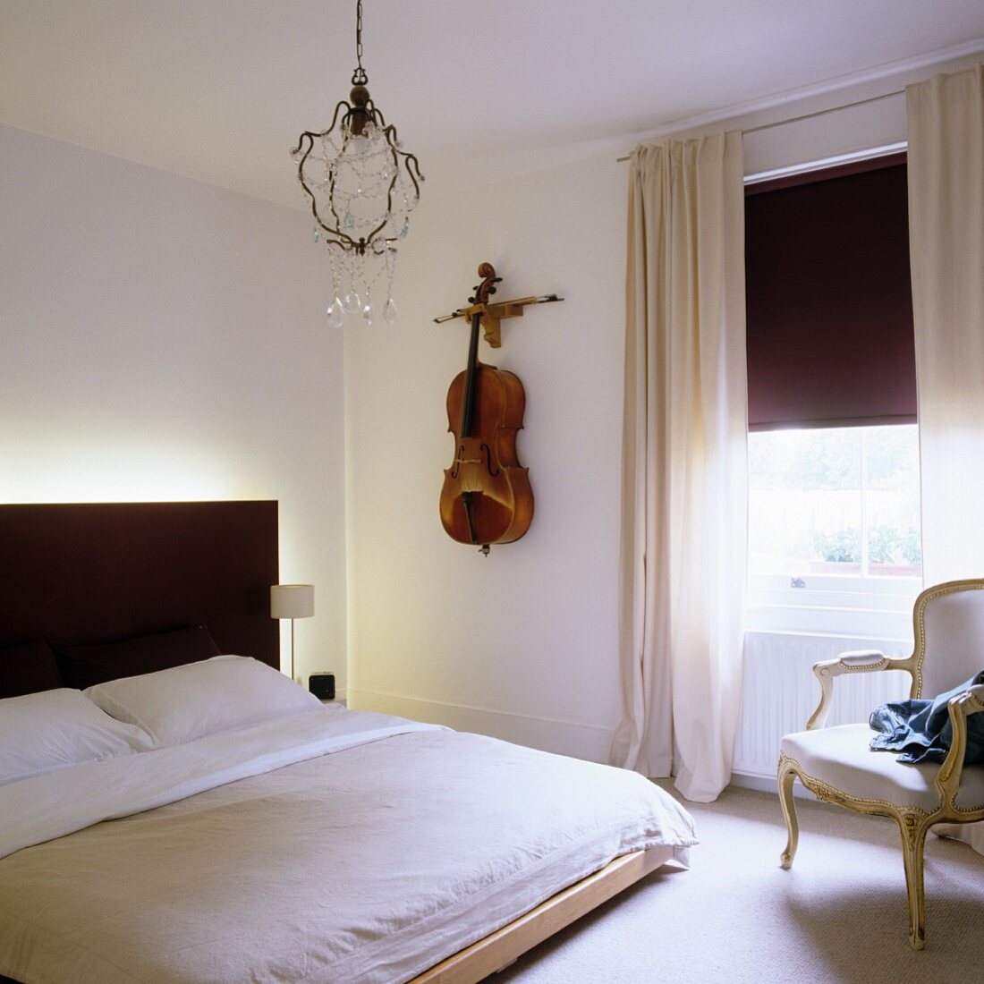 Modernes Bett mit schwarzem, beleuchtetem Kopfteil, Cello als Wanddeko neben Fenster mit halb geschlossenem Rollo
