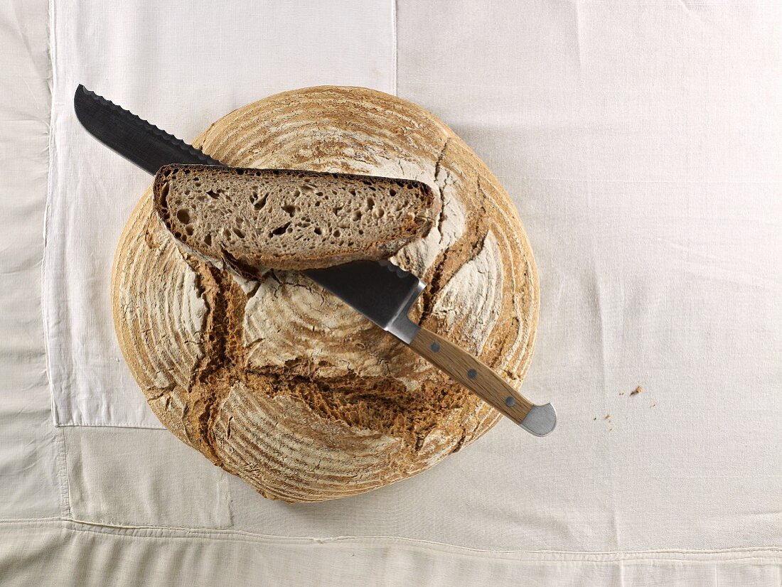 A brown loaf, with a bread knife and a slice of bread lying on top of it