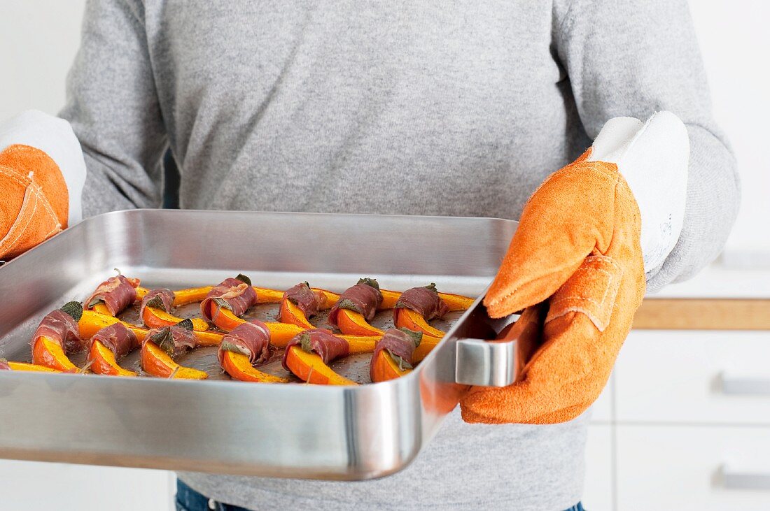 A man holding a roasting tin of squash slices wrapped in bacon with sage