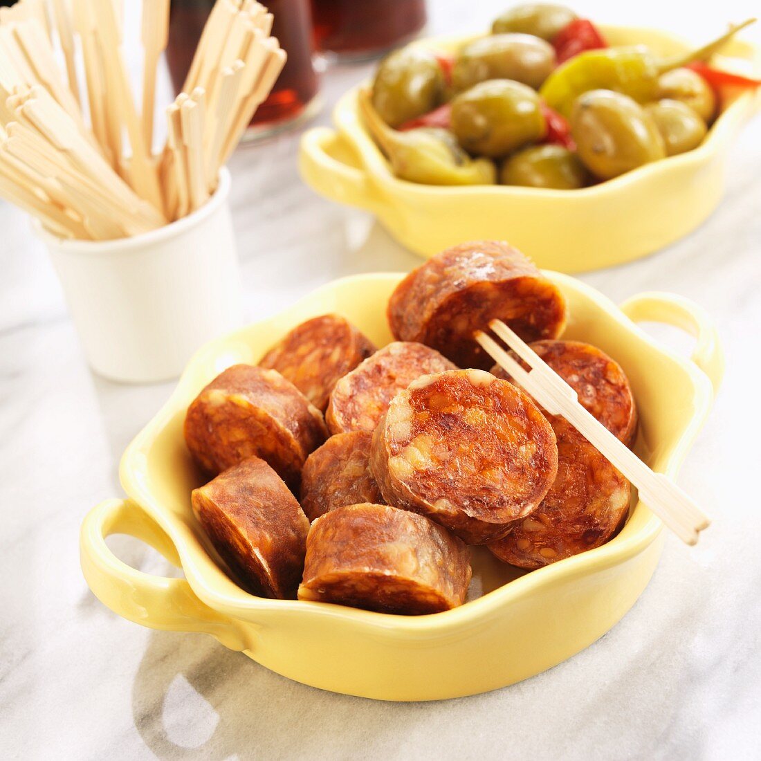 Tapas; Sliced Chorizo Sausage in Foreground, Olives and Peppers in Background; Toothpicks