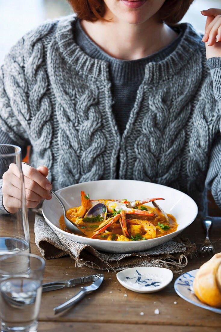 A woman in a grey woollen jumper eating bouillabaise at a wooden table laid with maritime decorations