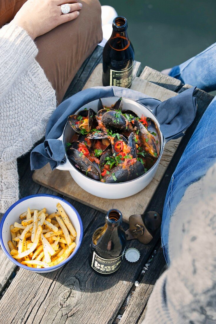 Ein Topf Miesmuscheln in Chili-Tomaten-Sud und Pommes auf Holzsteg zwischen Paar