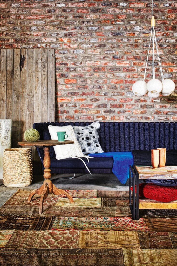 Rustic wooden side table on patchwork rug in front of dark blue sofa against rustic brick wall; pendant lamp with bundle of spherical lampshades