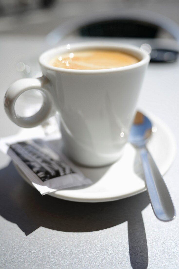 Milchkaffee in weißer Tasse, mit Zuckertütchen und Löffel