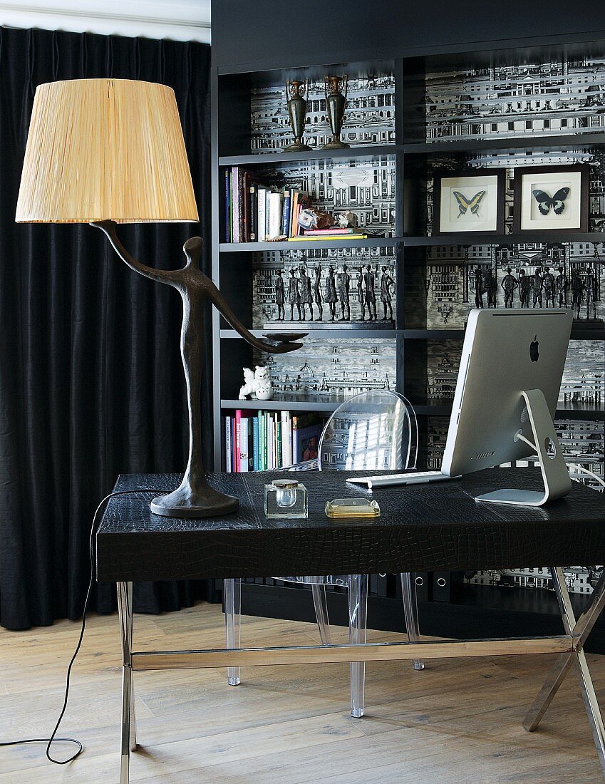 Table lamp with human figurine as base and computer on table in front of Ghost chair and black fitted shelving