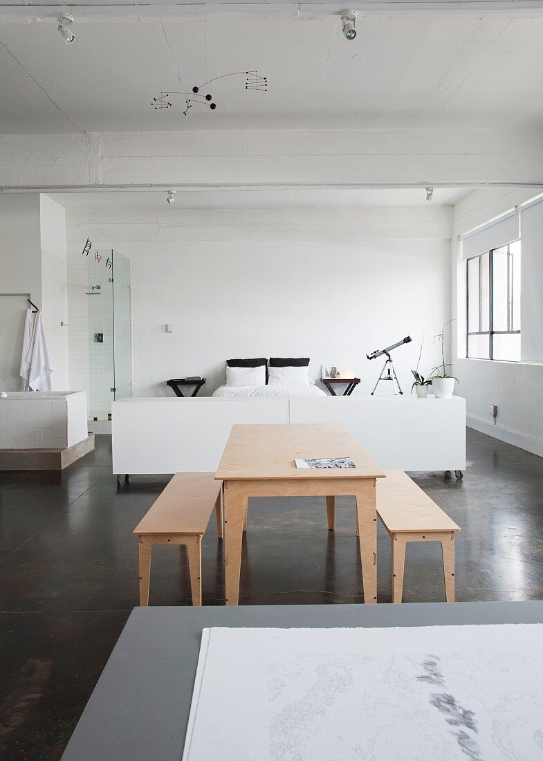 Pale wooden table and benches on grey concrete floor in front of white sleeping area in loft-style interior