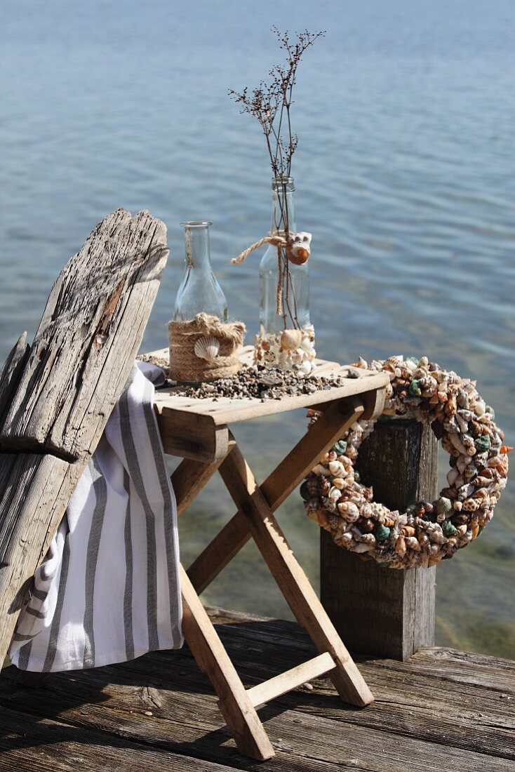 Mit Muscheln und Jute-Seilzopf verzierte Flaschen auf einem Klapptisch und Muschelkranz auf Holzsteg am Seeufer