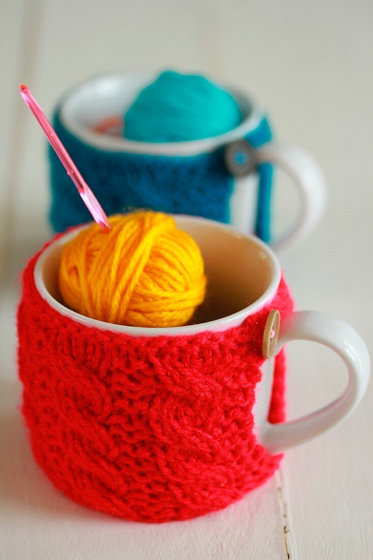 Mugs with knitted covers, balls of wool and crochet needle