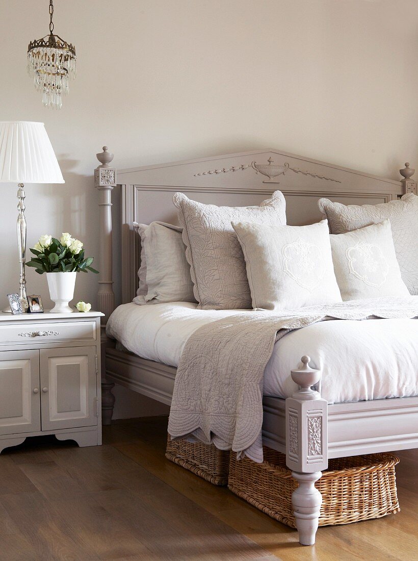Pale grey antique bed with headboard and carved ornamentation next to bedside table and table lamp