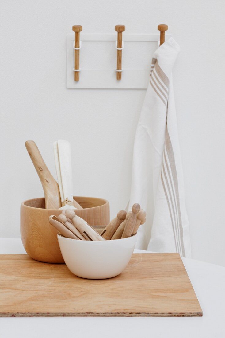 Kitchen utensils in white and wooden bowls and chopping board on table in front of tea towel hanging from hook rack