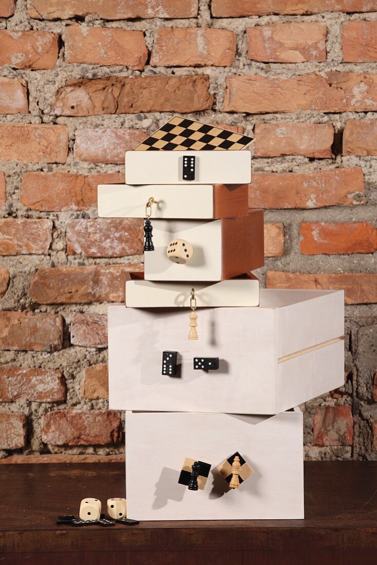 Gestapelte Kisten in Weiß mit angeschraubten Brettspielfiguren und Würfeln vor rustikaler Ziegelwand