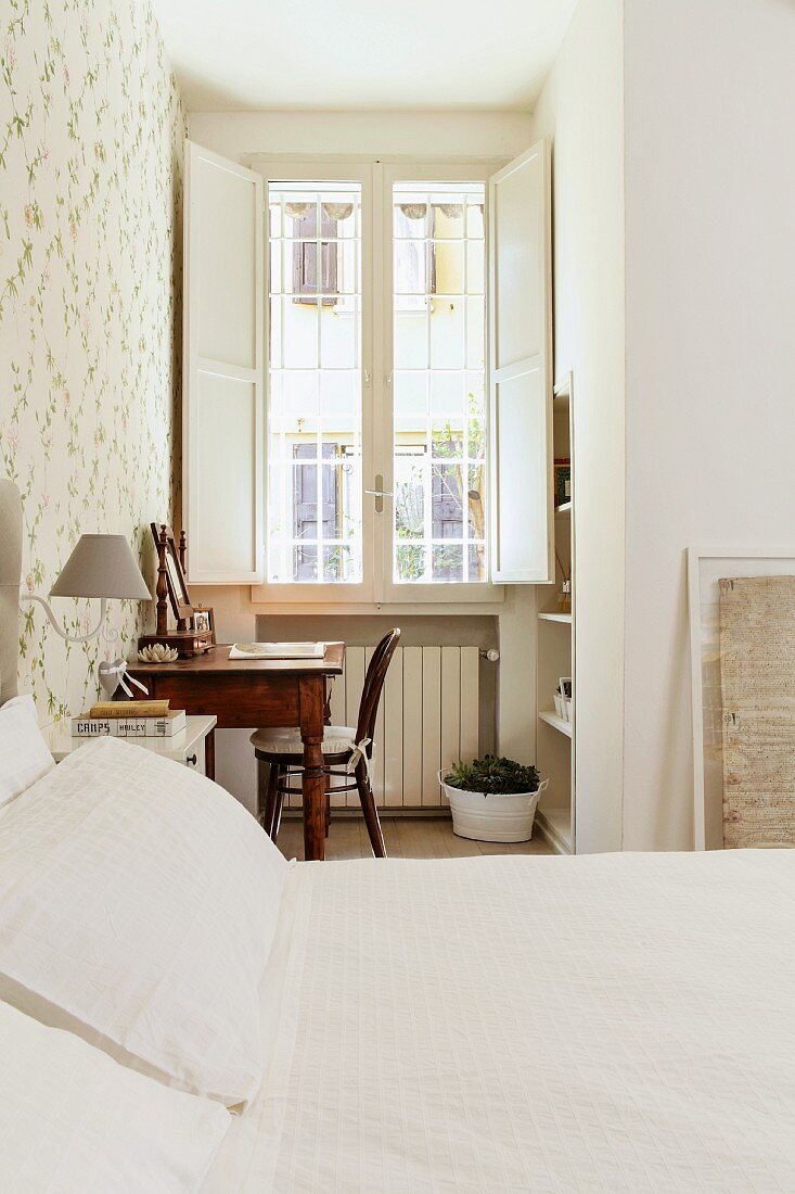 Blick über Doppelbett mit heller Tagesdecke auf antiken Schreibtisch mit Stuhl vor Fenster mit geöffneten Läden