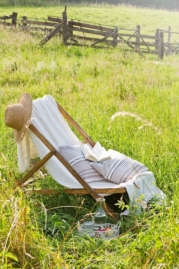 Liegestuhl mit Strohhut, Kissen und Buch auf einer Sommerwiese