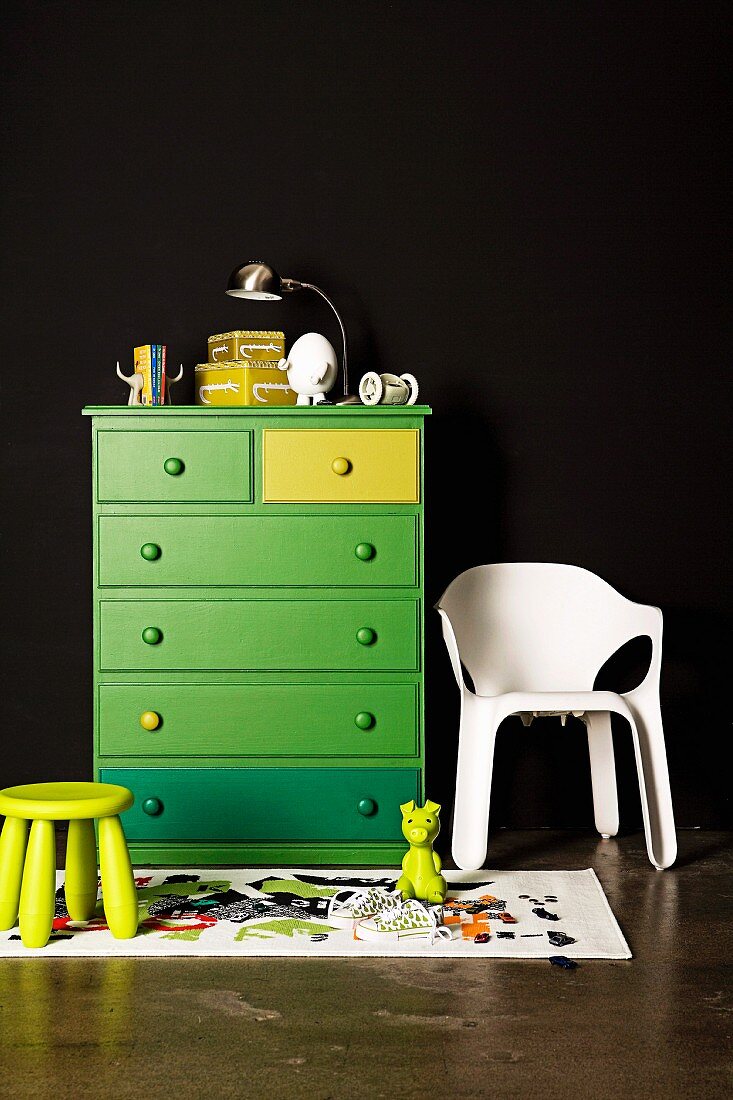 Green chest of drawers on black wall; next to it a white plastic chair and a children's stool