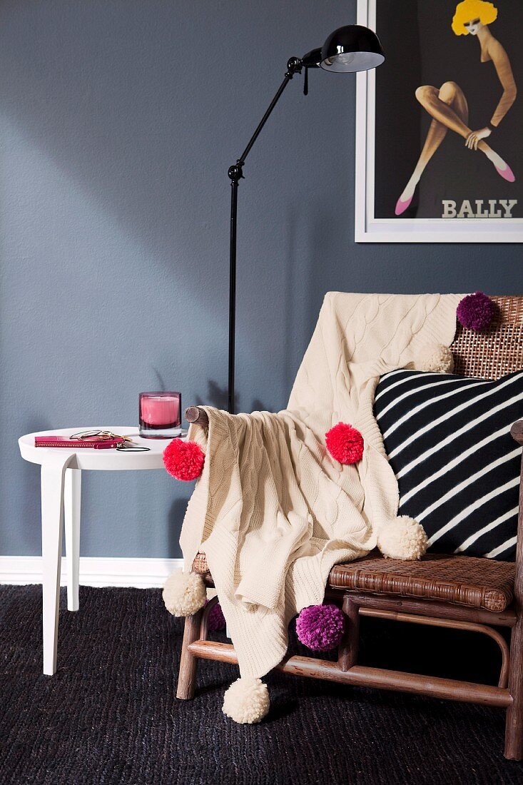 Pale, knitted blanket decorated with colourful wool pompoms on wicker chair