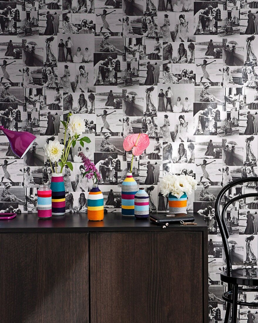 Wallpaper with pattern of black and white photos behind bottles wrapped in colourful stripes on 70s sideboard