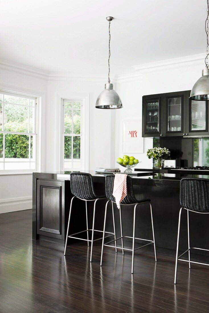 Elegant, black fitted kitchen with counter and bar stools on fine, dark parquet floor; white bay window and chrome pendant lamps