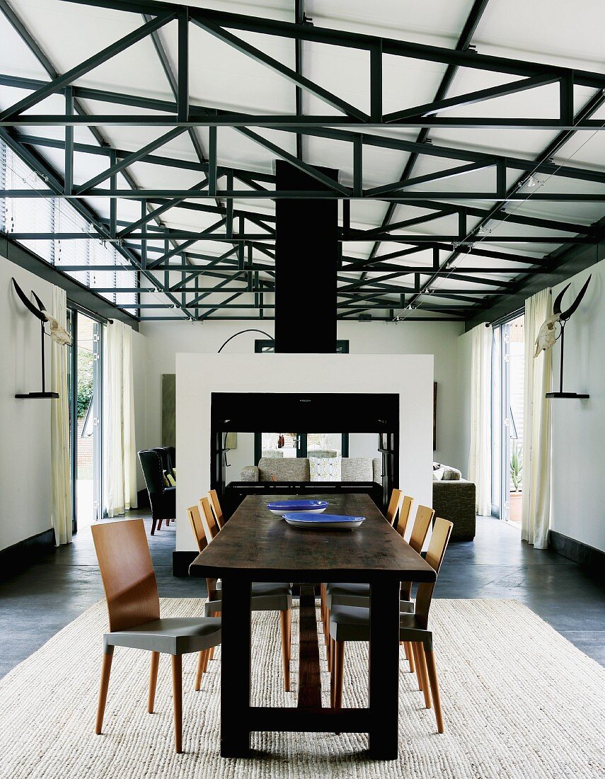 Rustic, dark wood dining table and retro-style chairs on rug in hall-style interior with exposed roof structure; partition in front of lounge area