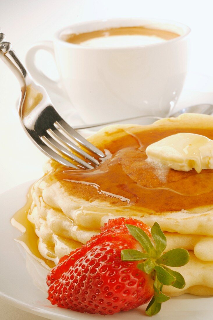 Gabel sticht in Pancakes mit Butter und Ahornsirup vor Kaffeetasse