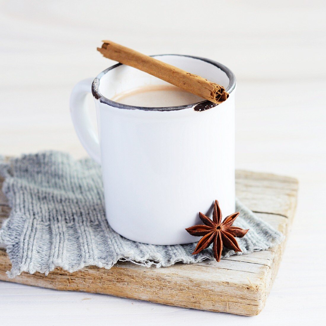weiße Emaille Tasse mit Tee, darauf liegt eine Zimtstange und ein Anisstern lehnt an der Tasse, alles steht auf einem gestrickten grauen Flicken und einem verwittertem Holzbrett
