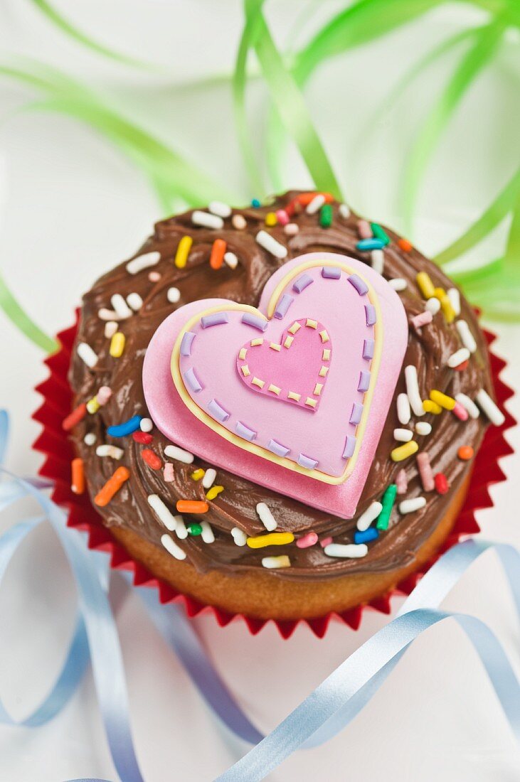 Cupcake mit Schokoladencreme, Zuckerstreuseln und rosa Herz zwischen Luftschlangen