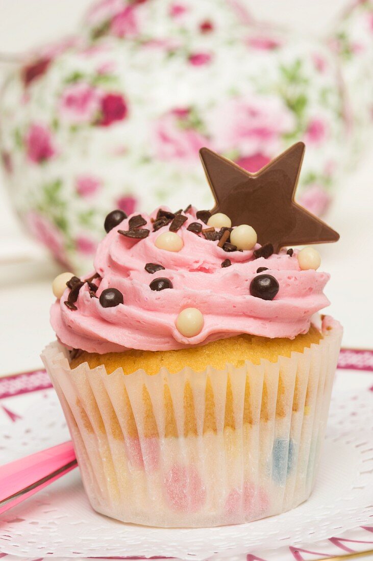 A pink cupcake decorated with a chocolate star in front of a teapot
