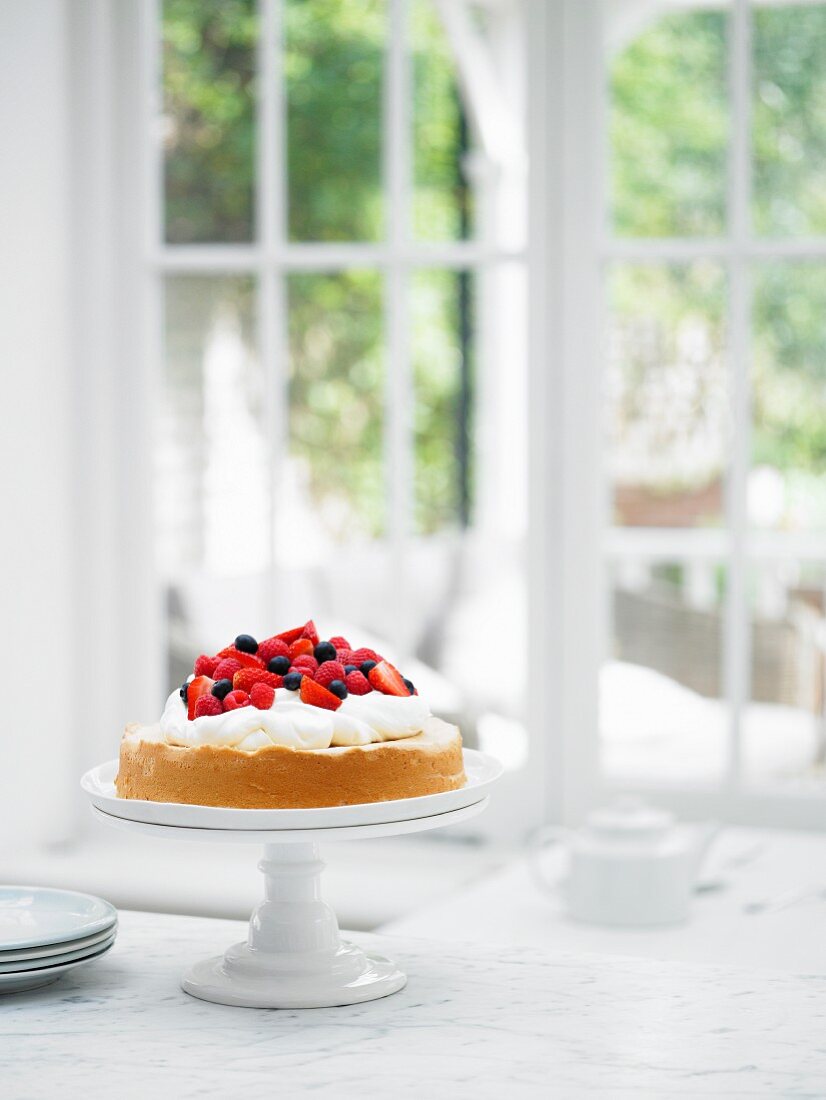 Sponge cake with cream and berries on a cake stand in the windos