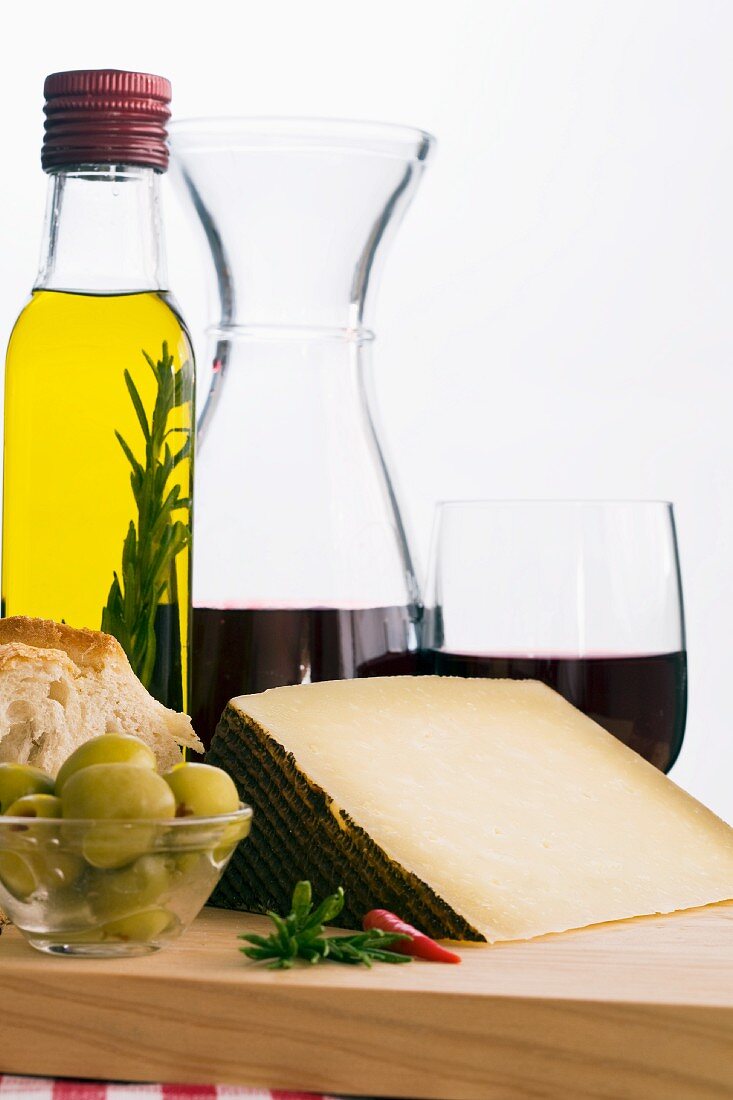 A still life featuring olive oil, red wine, bread, olives and Manchego (Spain)
