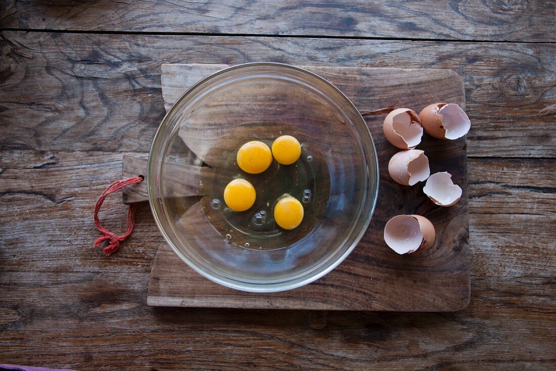 Aufgeschlagene Eier in Glasschüssel und Eierschalen auf Holzbrett