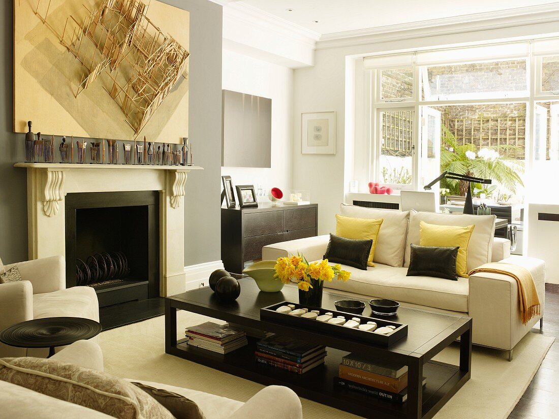 Pale sofa set and dark, box-shaped coffee table in elegant interior with wooden model suspended on wall and figurines on mantelpiece