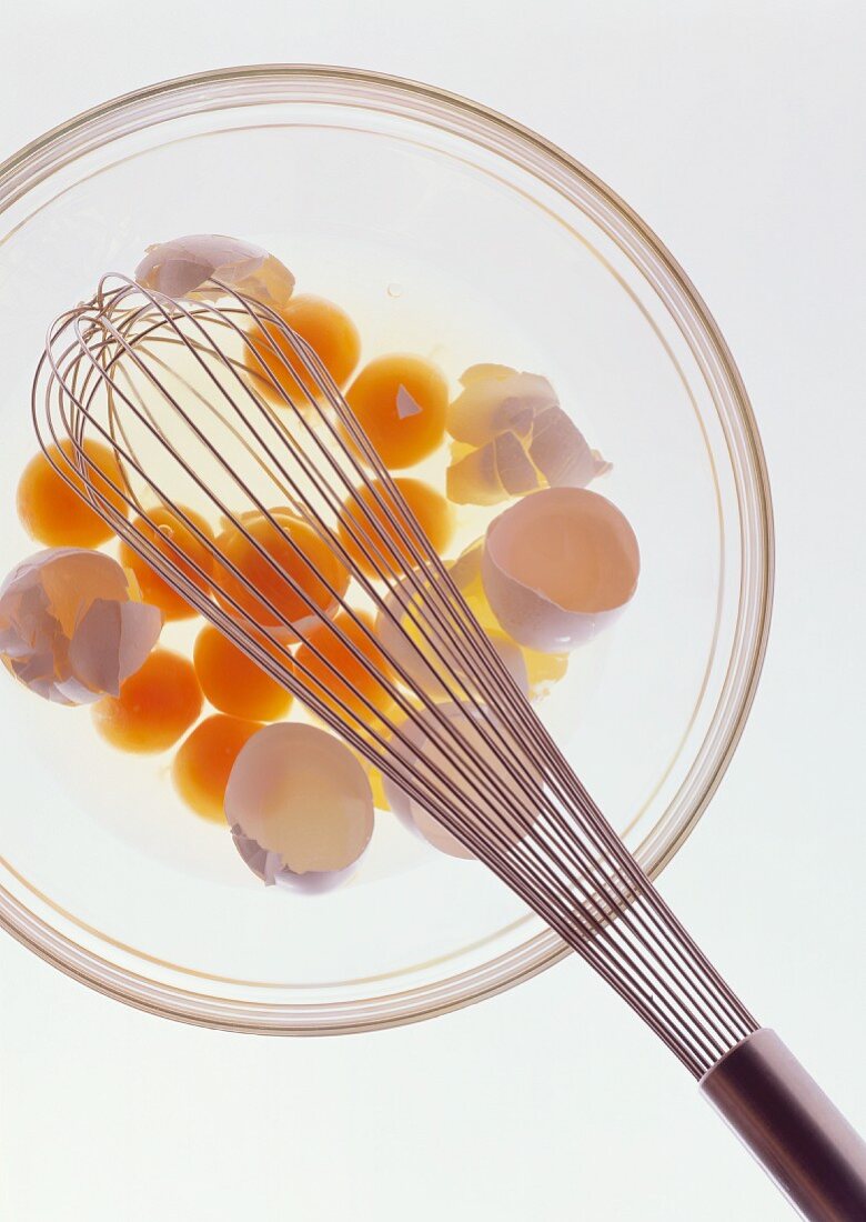 Glasschale mit aufgeschlagenen rohen Eiern, Eierschalen & Rührbesen