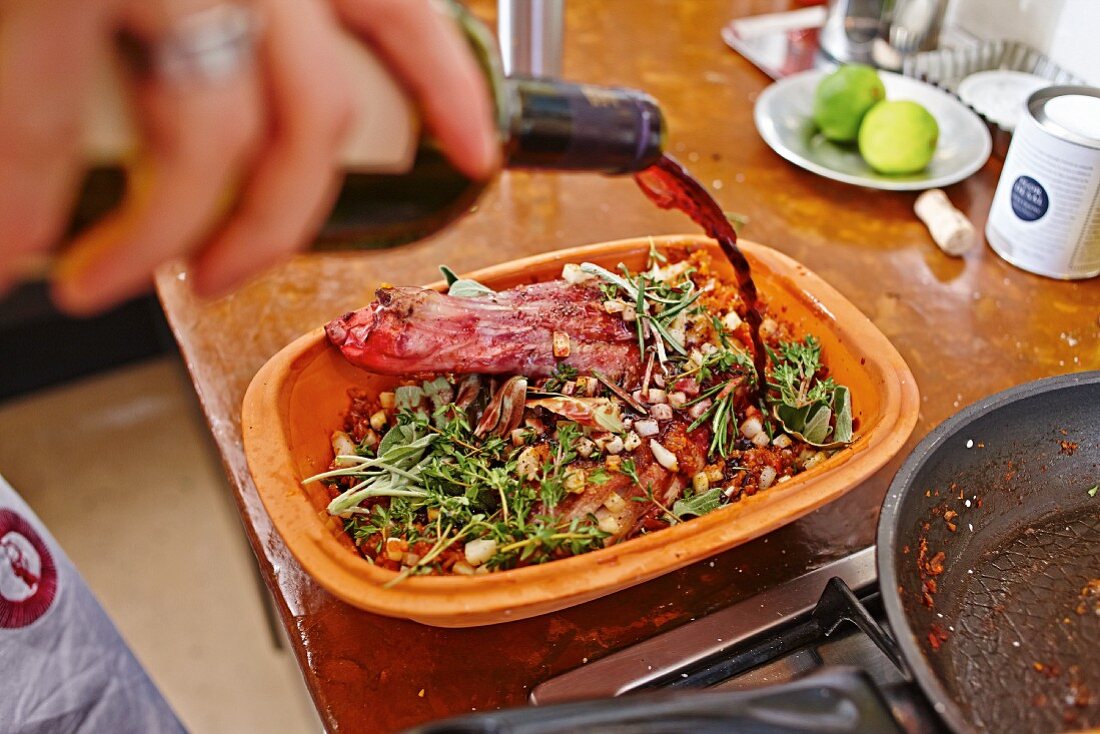 Red wine being poured over lamb shoulder in a clay roasting dish