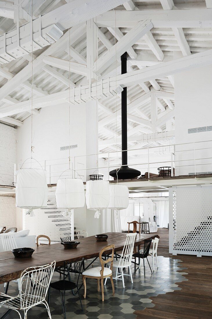 Dining table with various chairs on tiled floor area and pendant lamps with white fabric lamps in front of mezzanine in roof with whitewashed roof structure