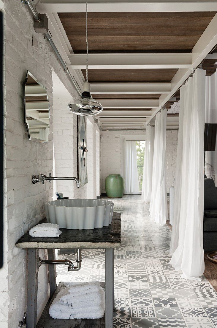 Wash basin on simple table with wall-mounted taps in tiled corridor area opposite open curtains used as partition