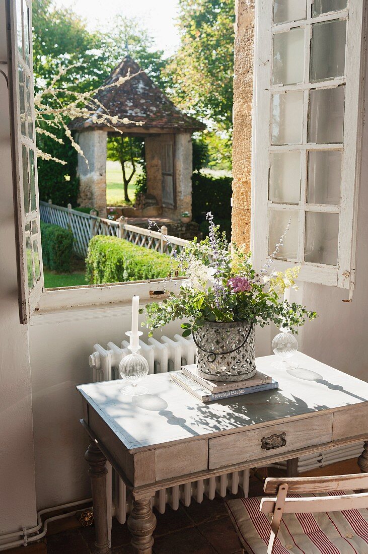 Bouquet of garden flowers on simple writing desk below open window with view of garden