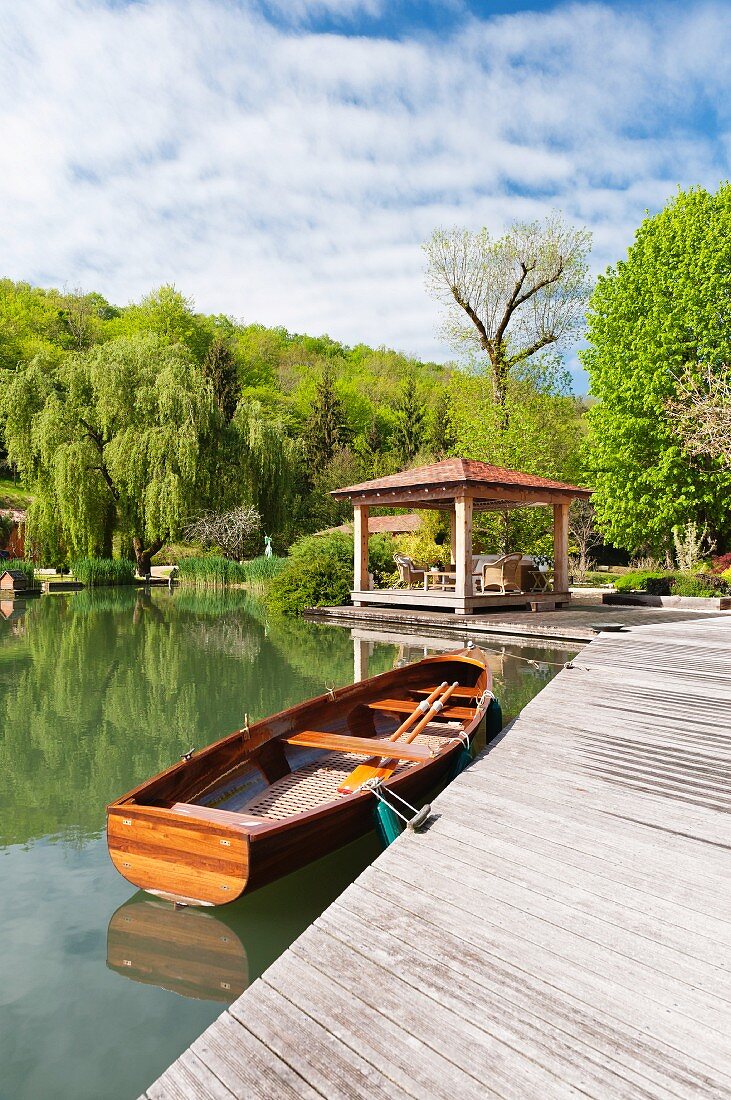 Ruderboot an einem Holzsteg mit Pavillon; bewaldeter Hügel im Hintergrund