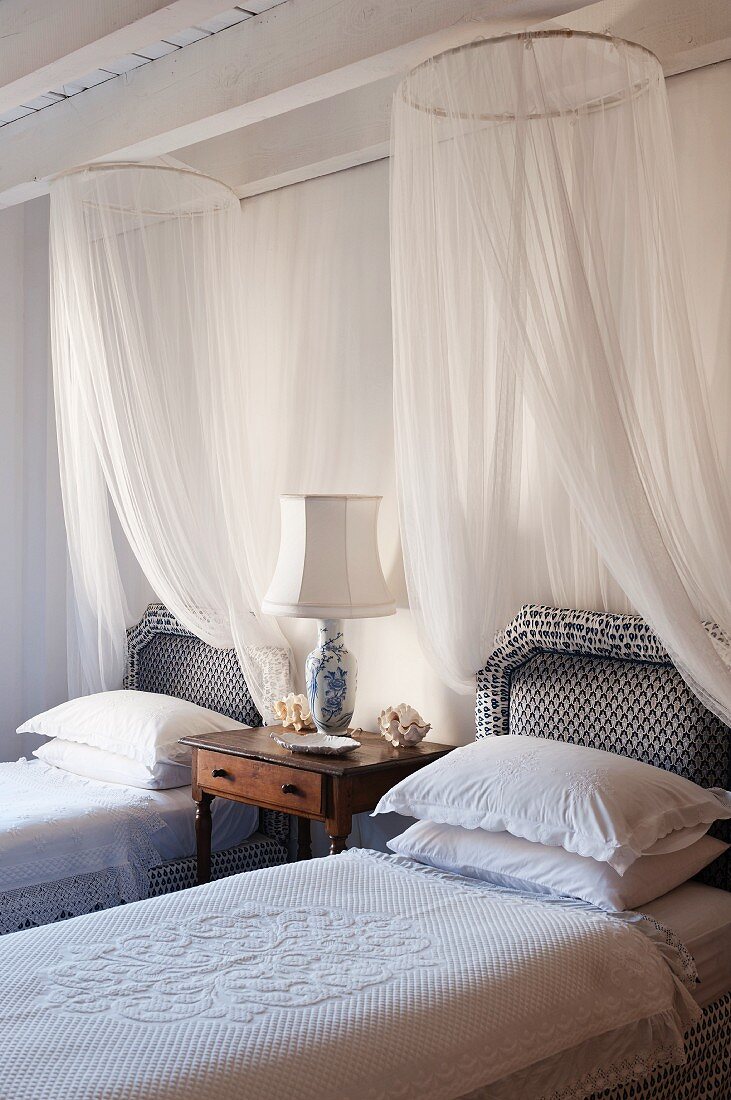 Two elegant, country-house-style twin beds with white mosquito nets either side of antique bedside table