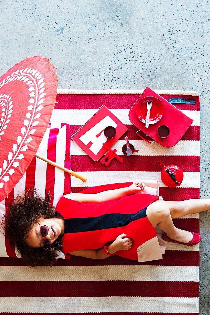 Red girls' accessories and toys on red and white striped rug; smiling girl in red dress lying on rug