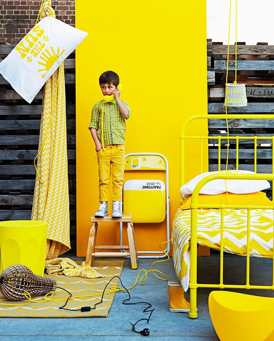 Yellow furnishings for a child's bedroom in front of yellow panel; small boy wearing yellow trousers standing on stool