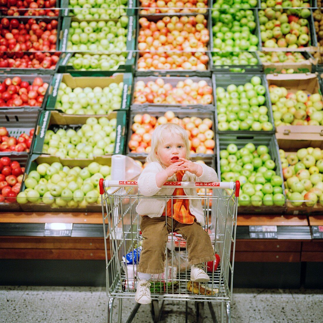 A small boy in a shopping trolley in front of a large display of apples
