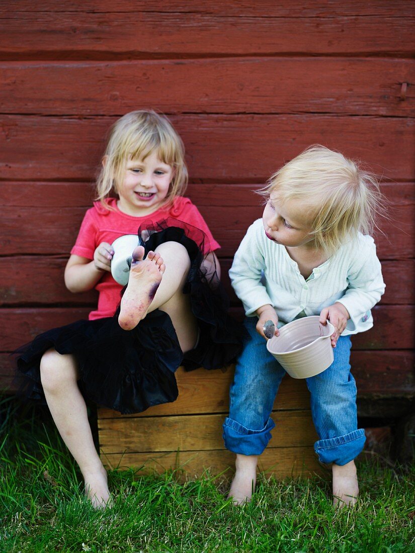 Little girls holding pots sitting and playing against outer wall of wooden cabin